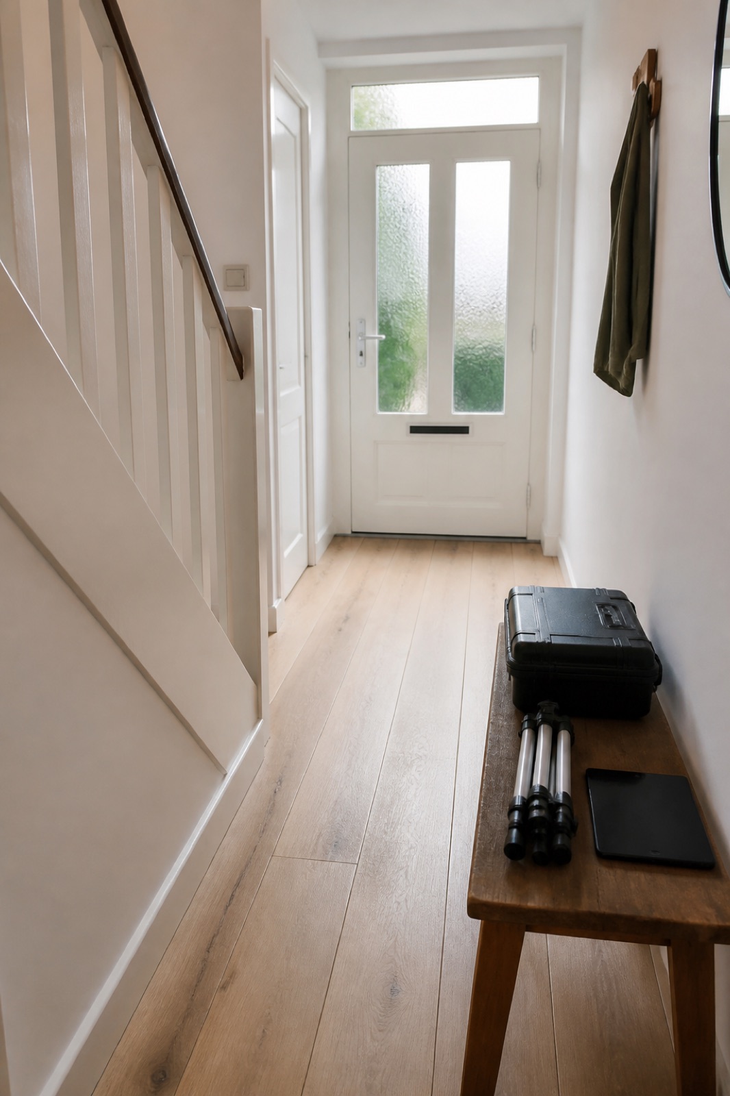 Lichte hal van een tussenwoning met een witte trap. Op een houten bankje liggen een koffer, een statief en een tablet.